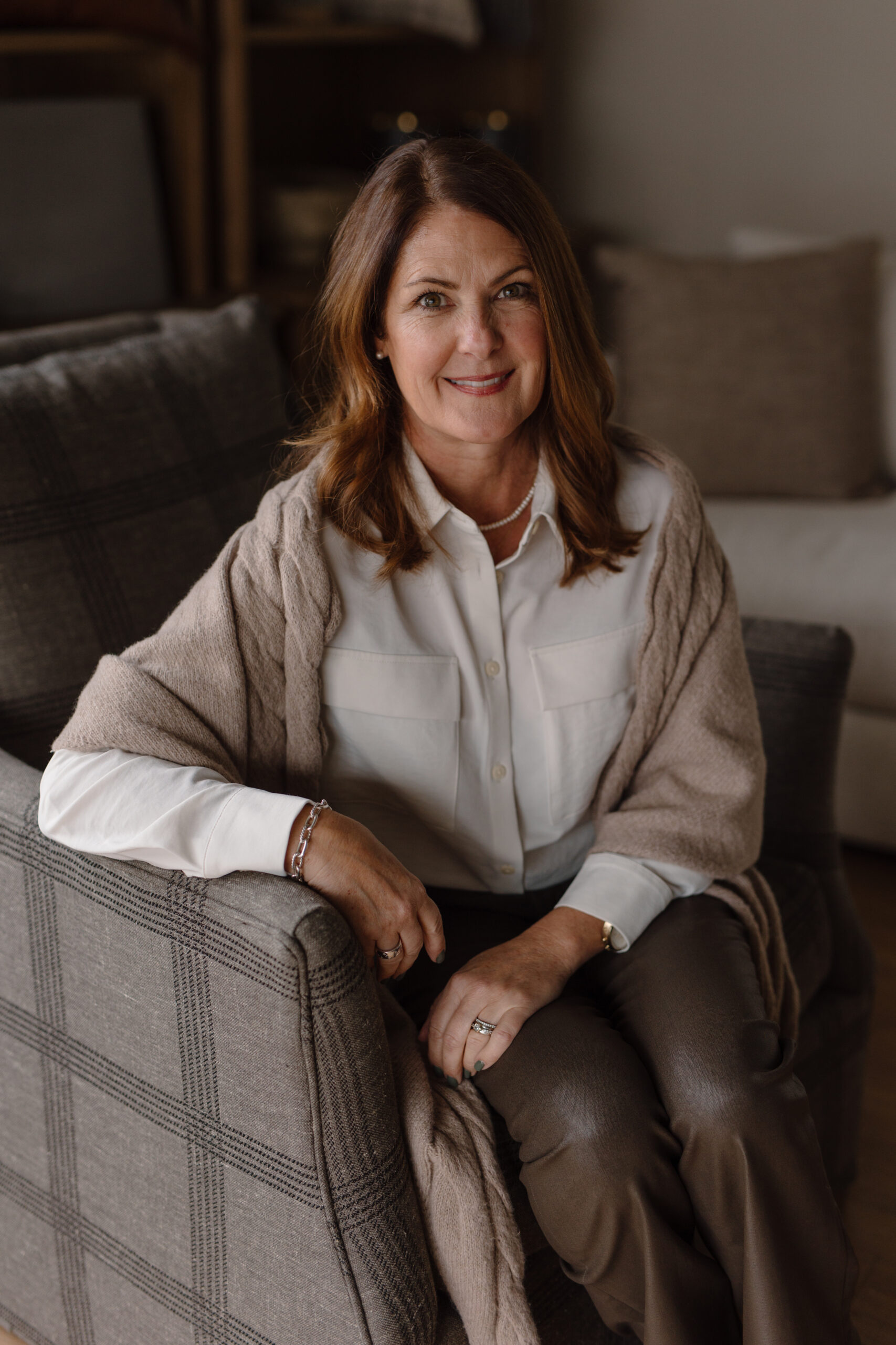 Corrie Witherall, an interior designer at Dwellings, sitting in a grey plaid armchair wearing a white collared shirt and a beige shawl.