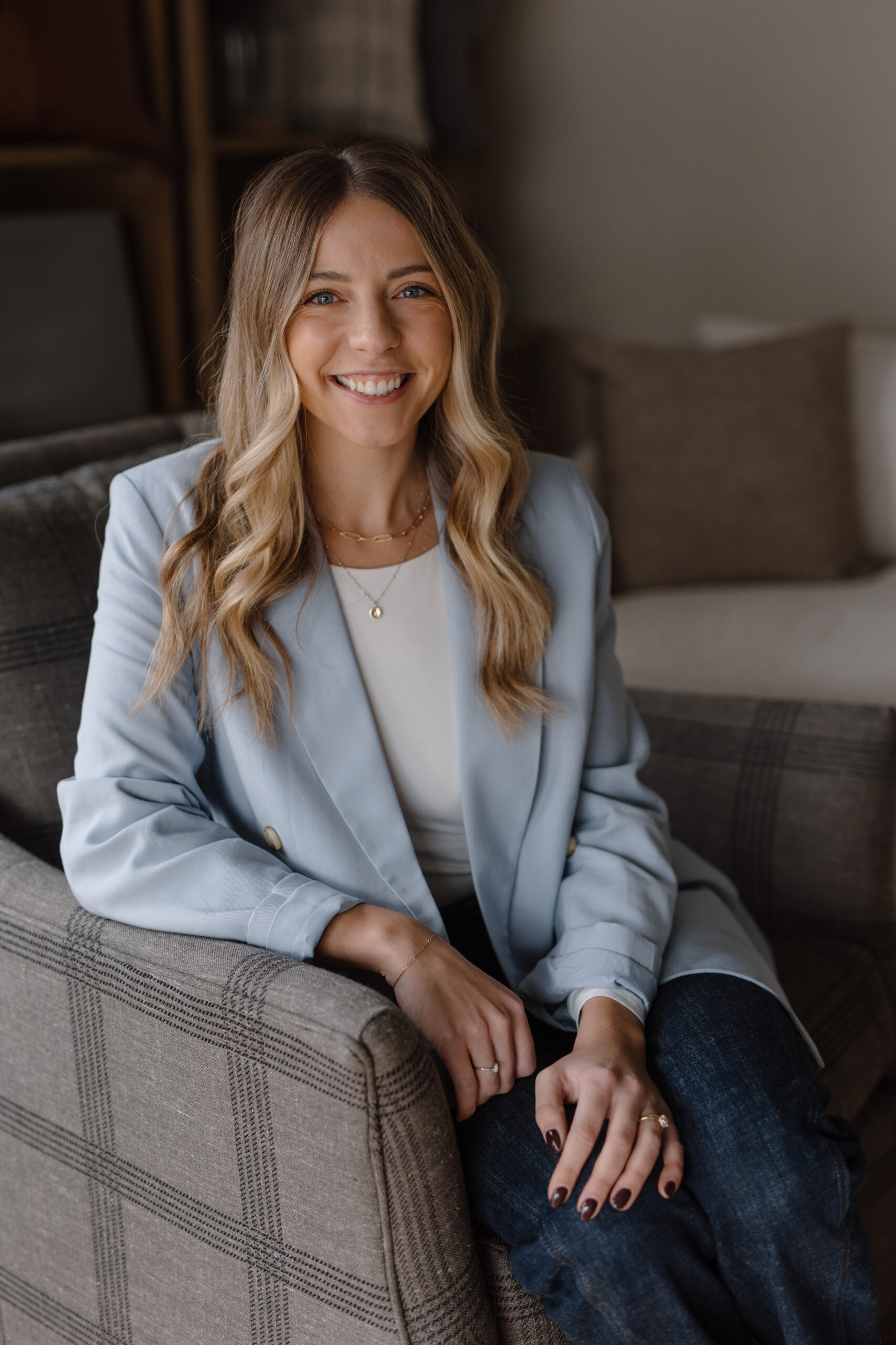 Emily Vander Boon | Founder & Designer at Emily Lynn Interiors Emily Vander Boon, designer and founder of Emily Lynn Interiors, wearing a light blue blazer and smiling while sitting in a grey plaid armchair.