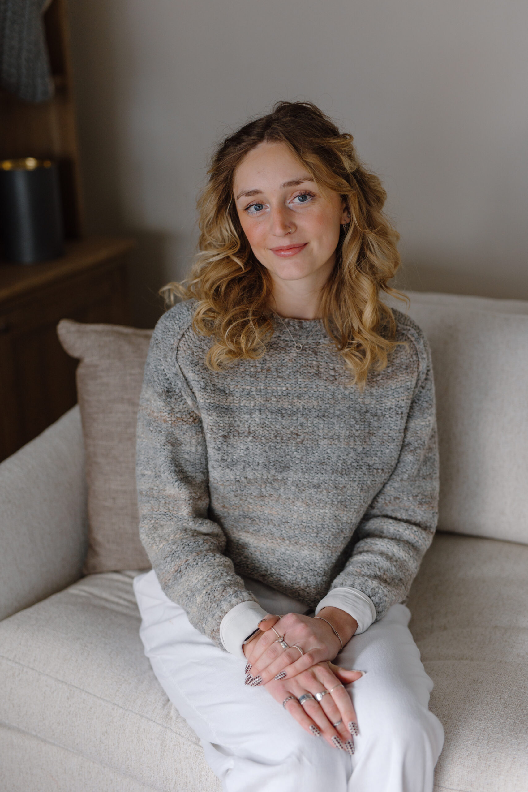 Sydney Eldred | Studio Manager at Dwellings Sydney Eldred, Studio Manager at Dwellings, sitting on a neutral sofa wearing a grey textured sweater and white trousers.